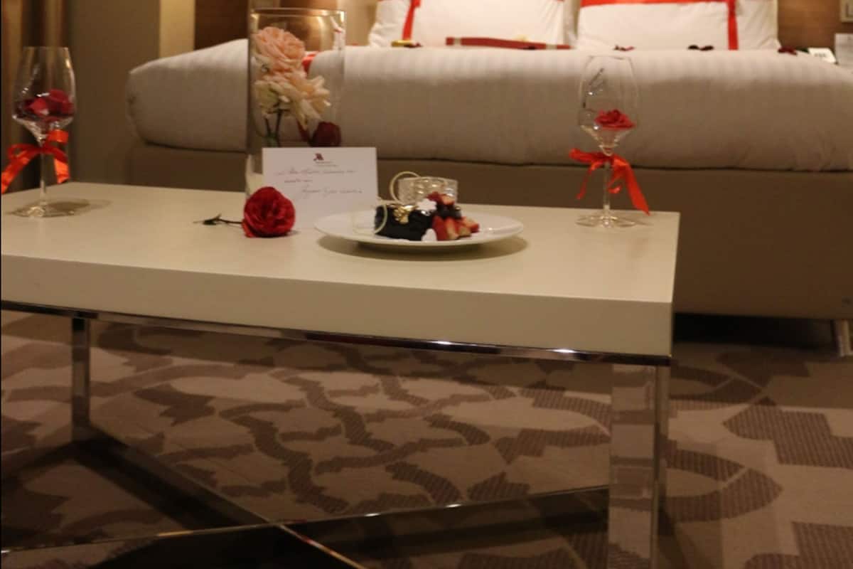 A hotel room with a neatly made bed, two bedside lamps, a table with a glass of wine and a rose, and red balloons with "I love you" written on them.