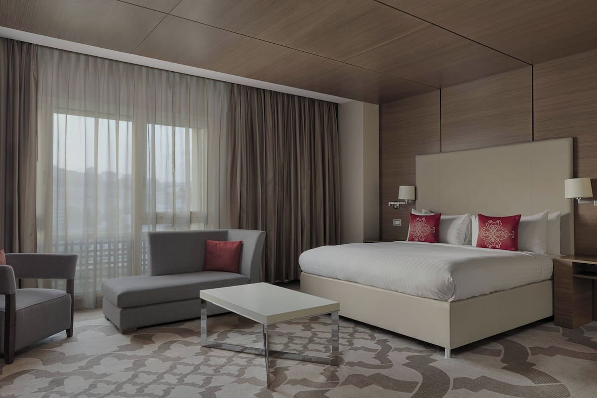 A modern hotel room with a large bed, a sofa, a small table, and a view of the city through large windows.