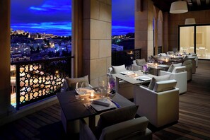 Restaurante no terraço com mesas preparadas para refeições, com vista para a paisagem urbana ao anoitecer.
