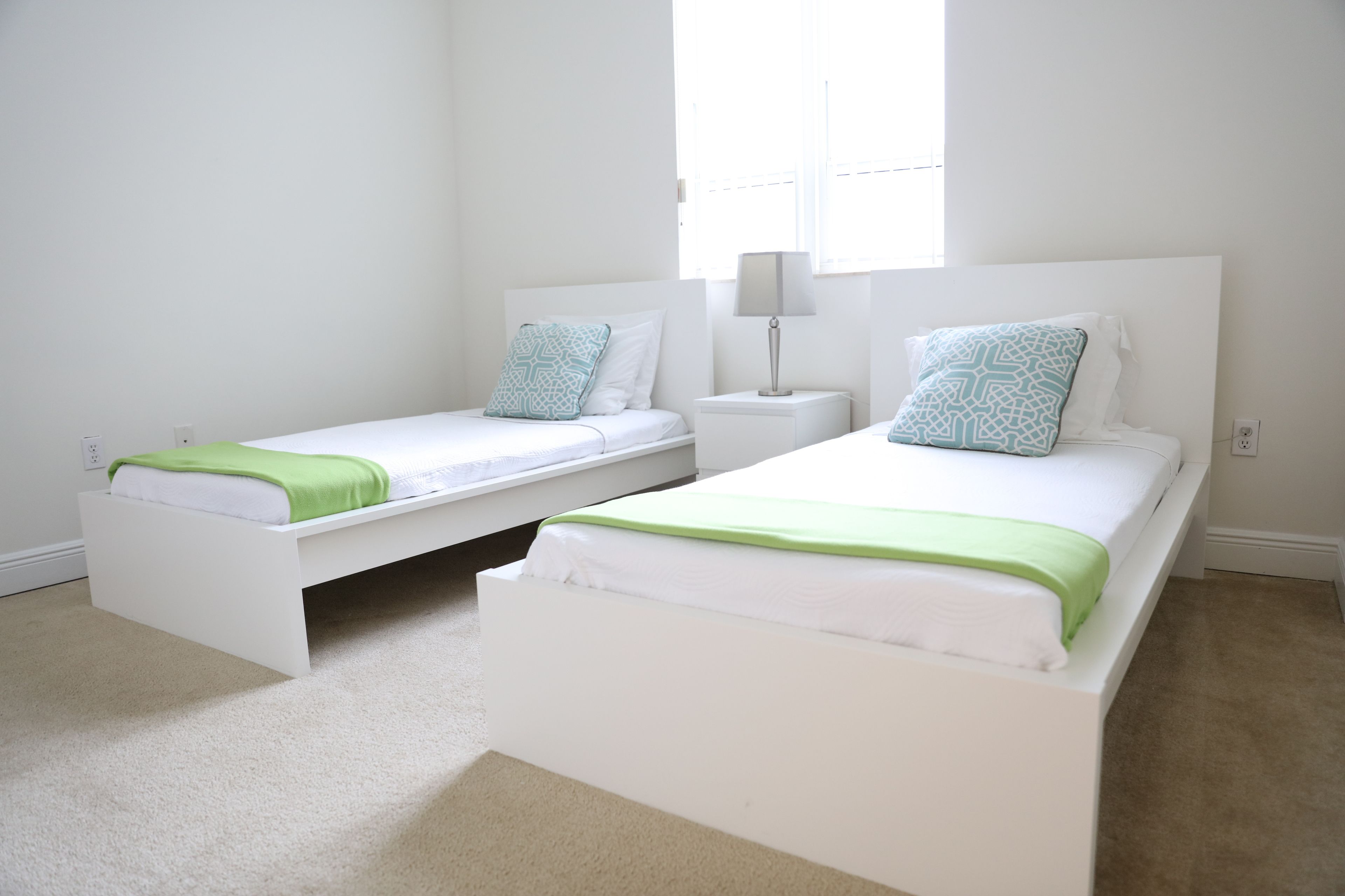 Two single beds with white frames and green bedding, a bedside table with a lamp, and a window providing natural light.