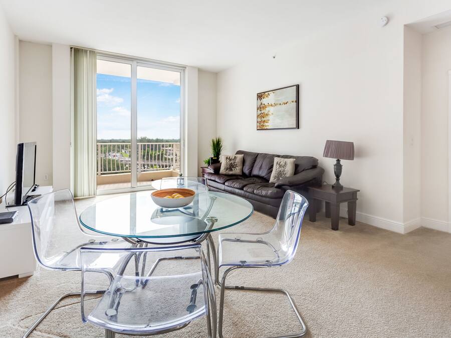A modern living room with a glass dining table, clear plastic chairs, a leather sofa, and a balcony with a view.