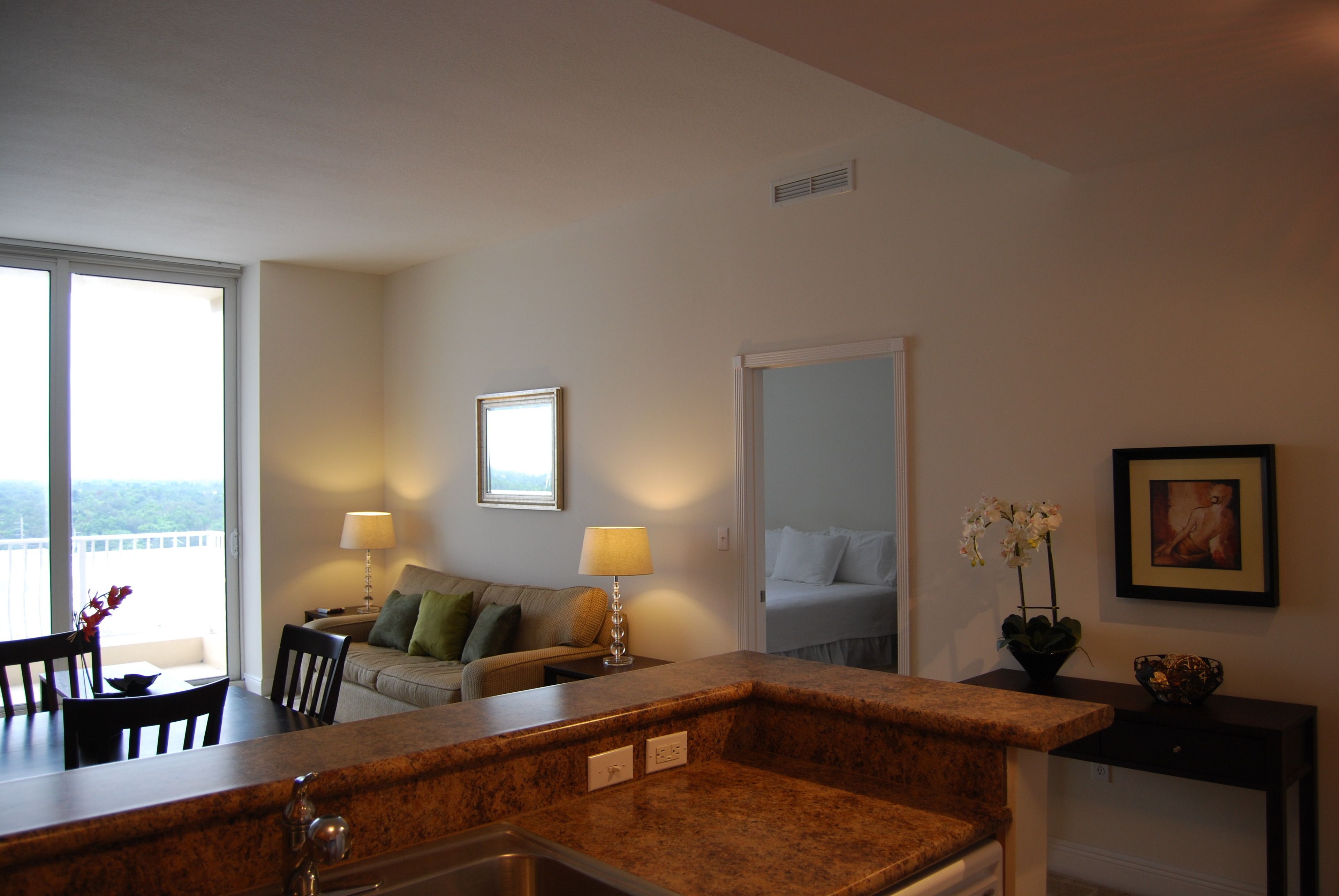 A modern kitchen with granite countertops, a dining area with a table and chairs, a sofa with a lamp, and a view through large windows.