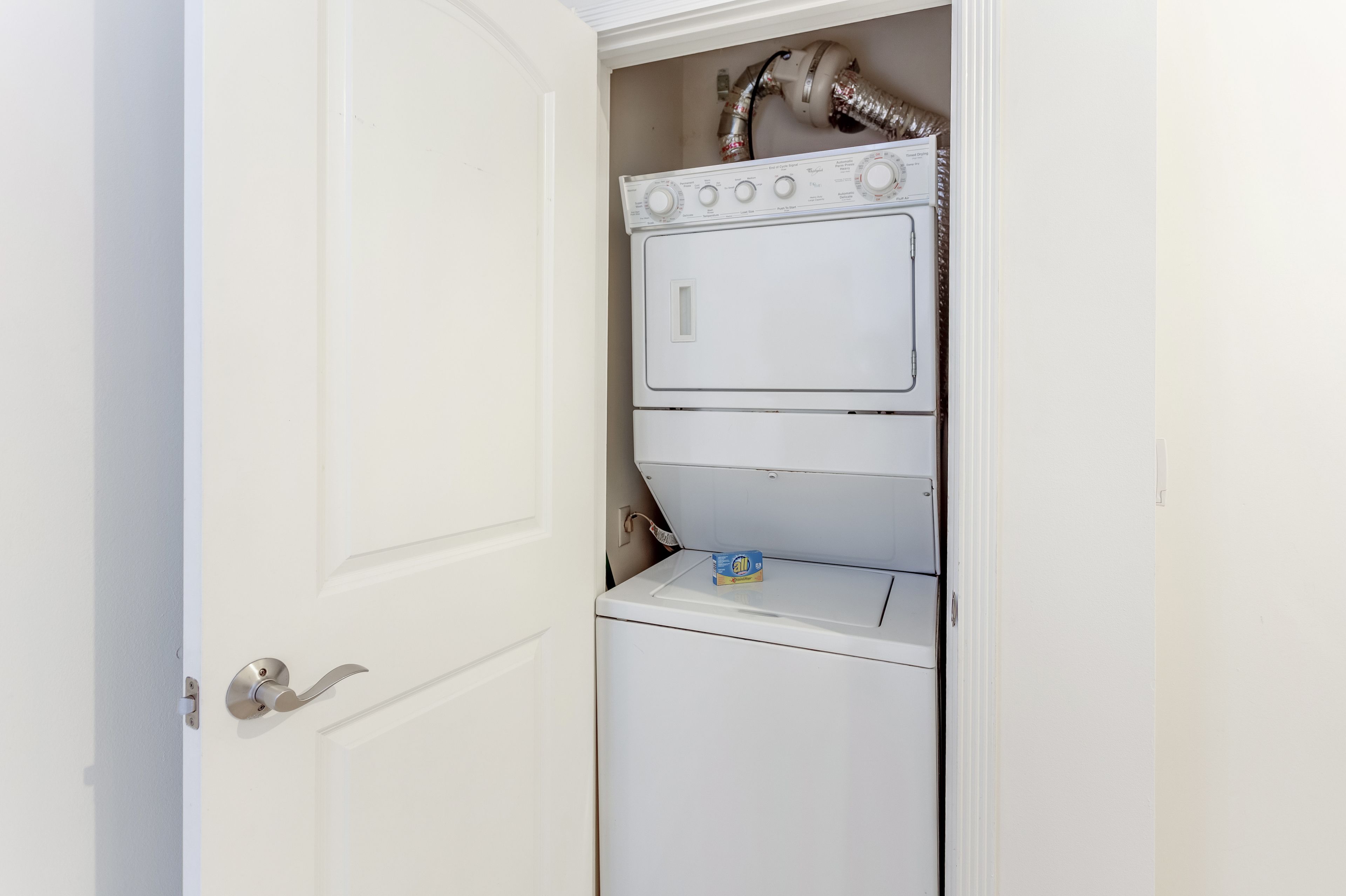 A compact laundry room with a stacked washer and dryer, a small shelf with a detergent box, and a visible water heater above the dryer.