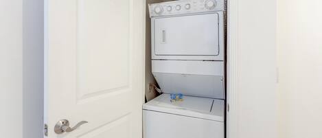 A compact laundry room with a stacked washer and dryer, a small shelf with a detergent box, and a visible water heater above the dryer.