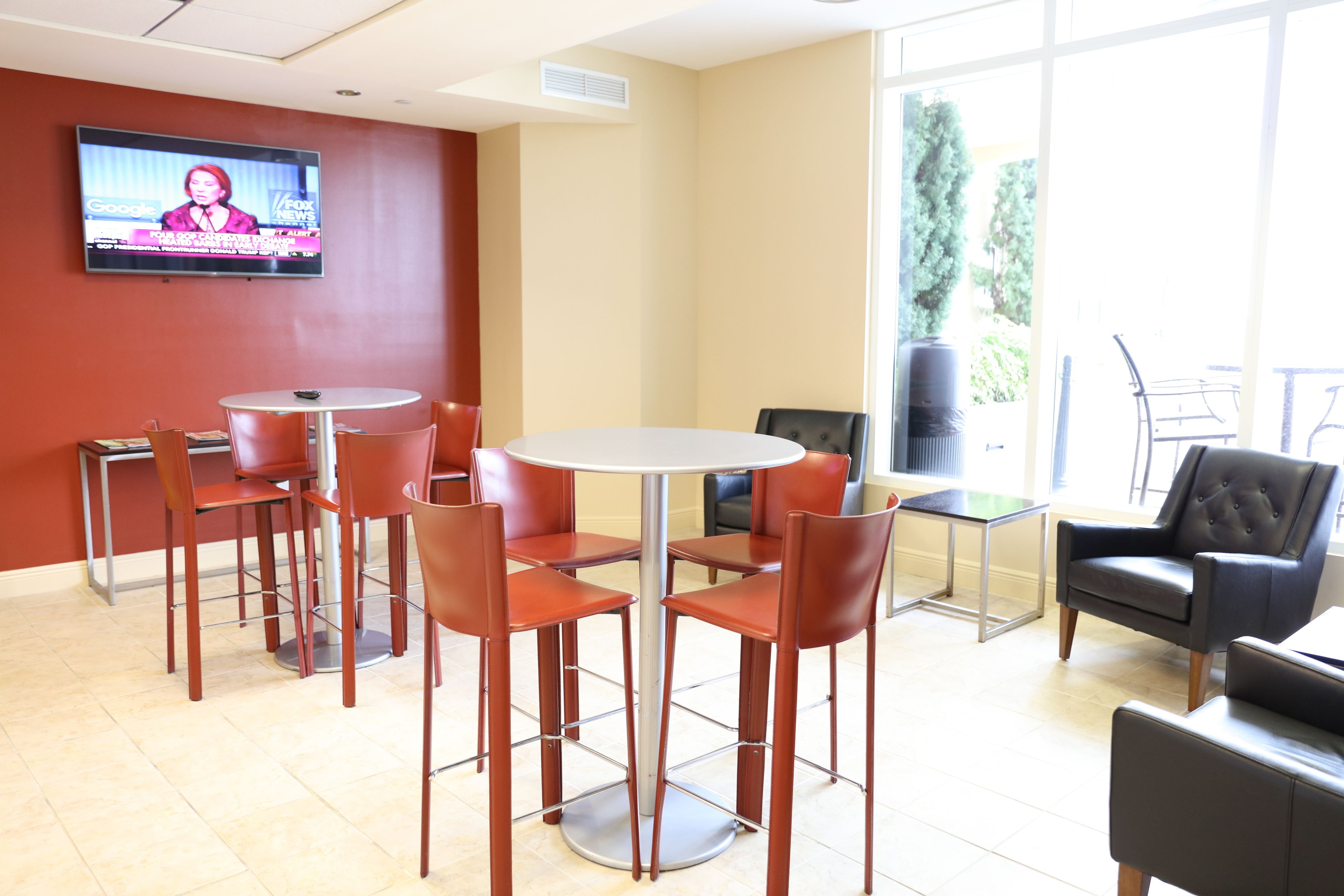 A modern conference room with a round table, red chairs, a television on the wall, and a view of the outdoors through large windows.