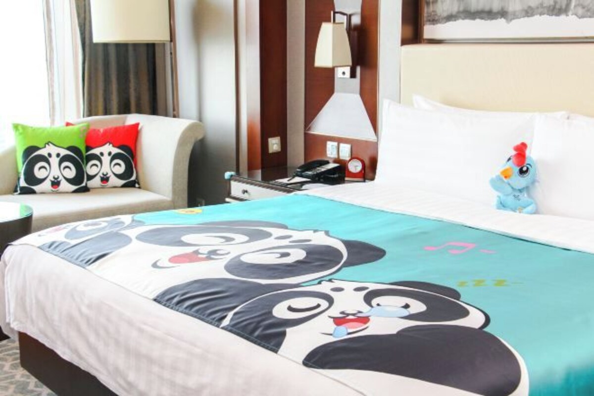 A hotel room with a bed featuring a panda-themed bedspread, a bedside table with a lamp, a chair with colorful cushions, and a small toy chicken on the bed.