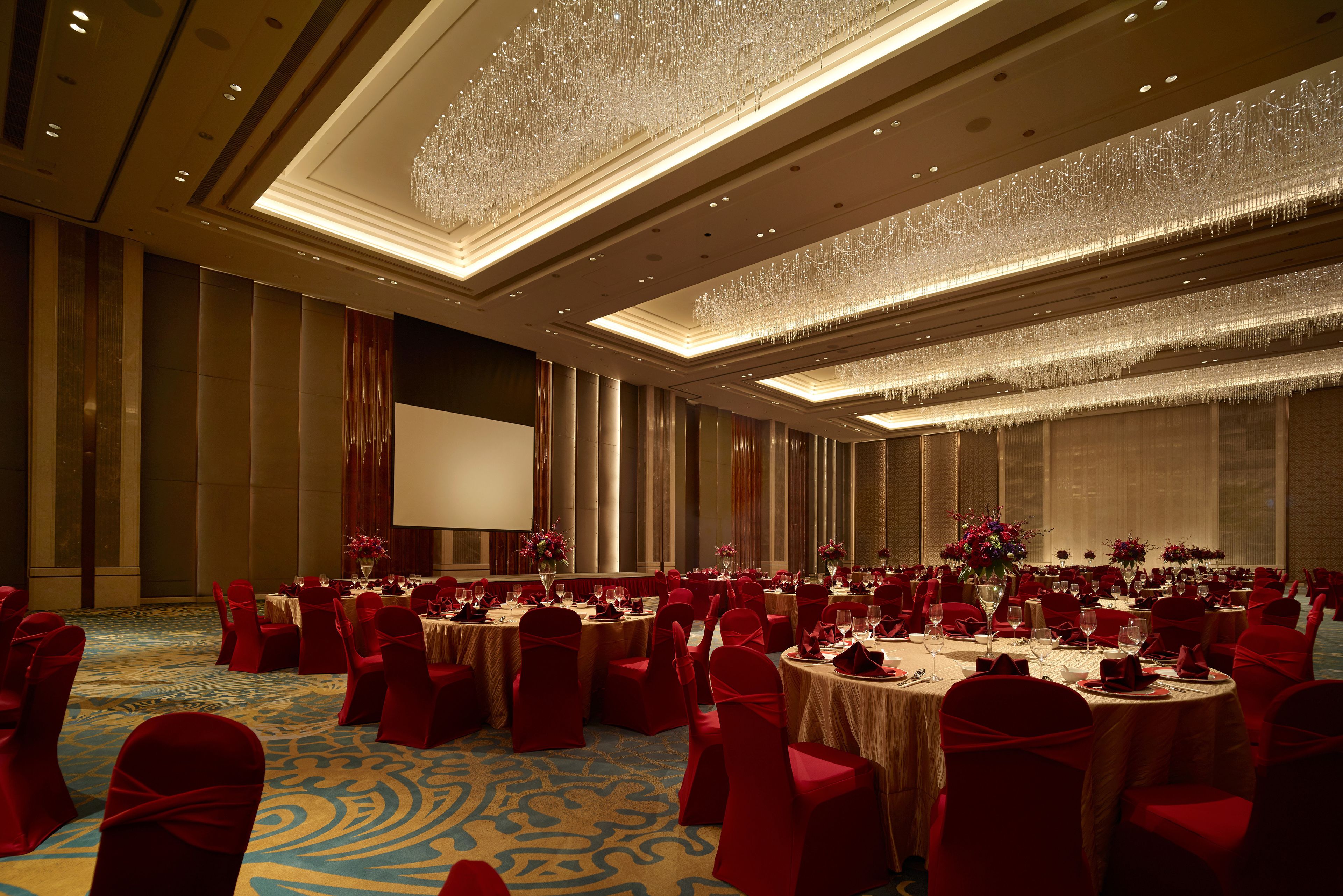 A banquet hall with round tables, red chairs, and a large screen.
