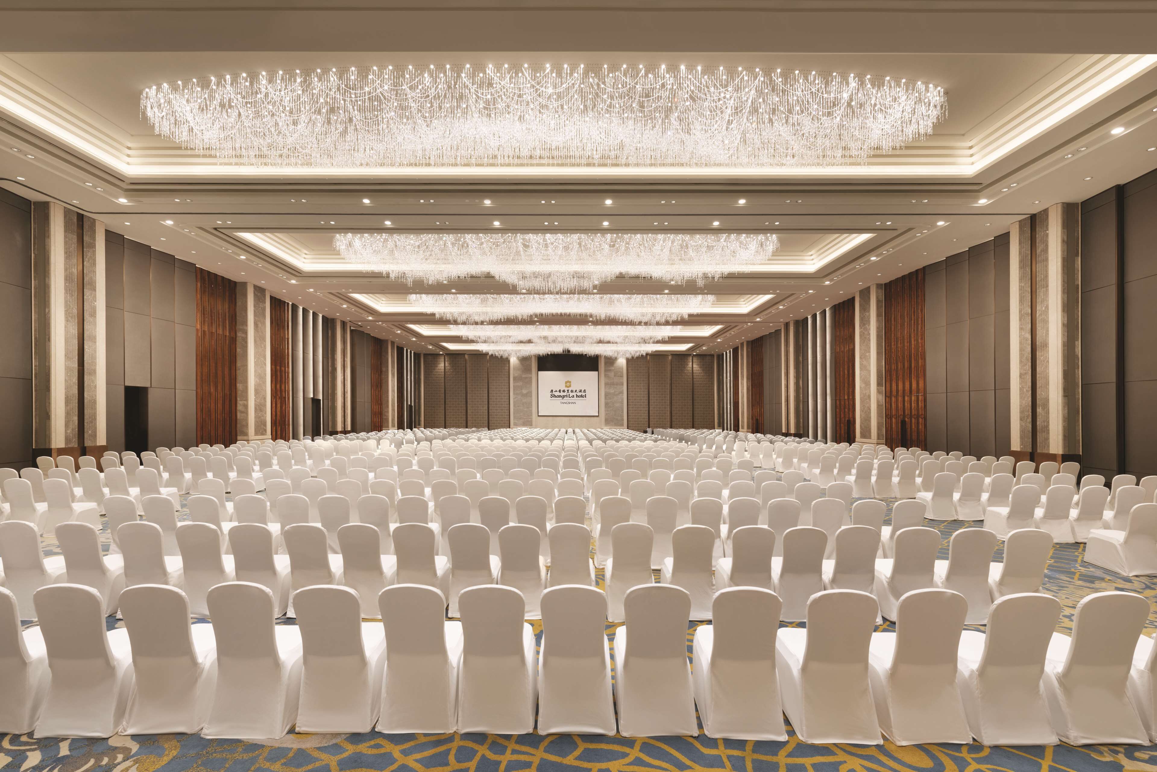 Une grande salle de conférence avec des rangées de chaises blanches, un lustre et un panneau sur le mur.