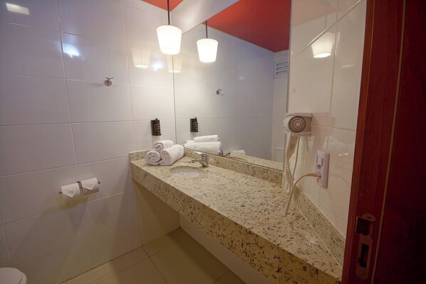 Un baño con azulejos blancos, encimera de granito, lavamanos, espejo, secador de pelo y toallas.
