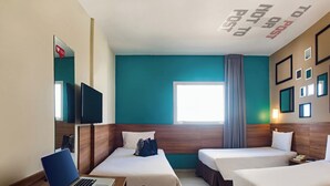 Une chambre d’hôtel avec deux lits, un bureau avec un ordinateur portable, une télévision et une fenêtre avec des rideaux.