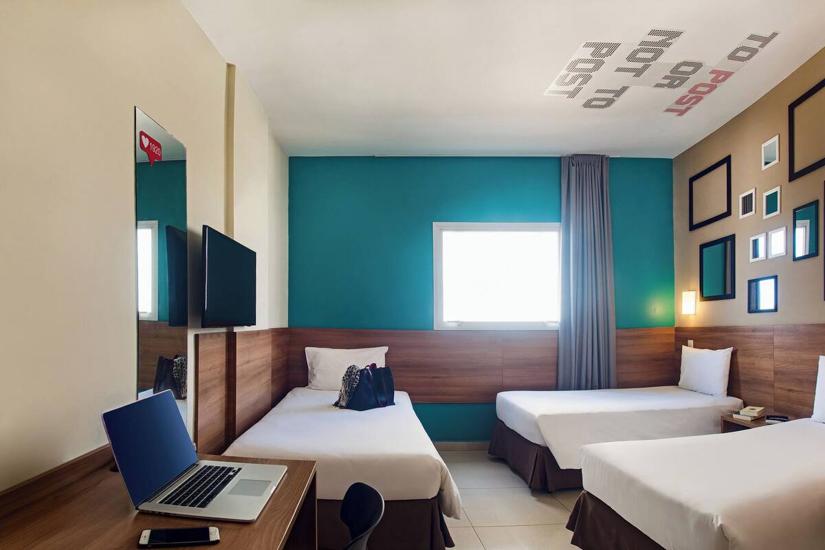 Een hotelkamer met twee bedden, een bureau met een laptop, een televisie en een raam met gordijnen.