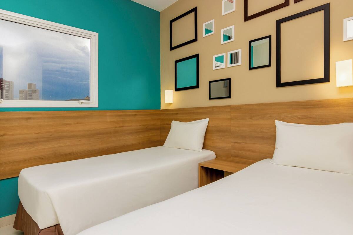 Een hotelkamer met twee eenpersoonsbedden, een houten hoofdbord, muur in blauwgroen en een raam met uitzicht op de stad.