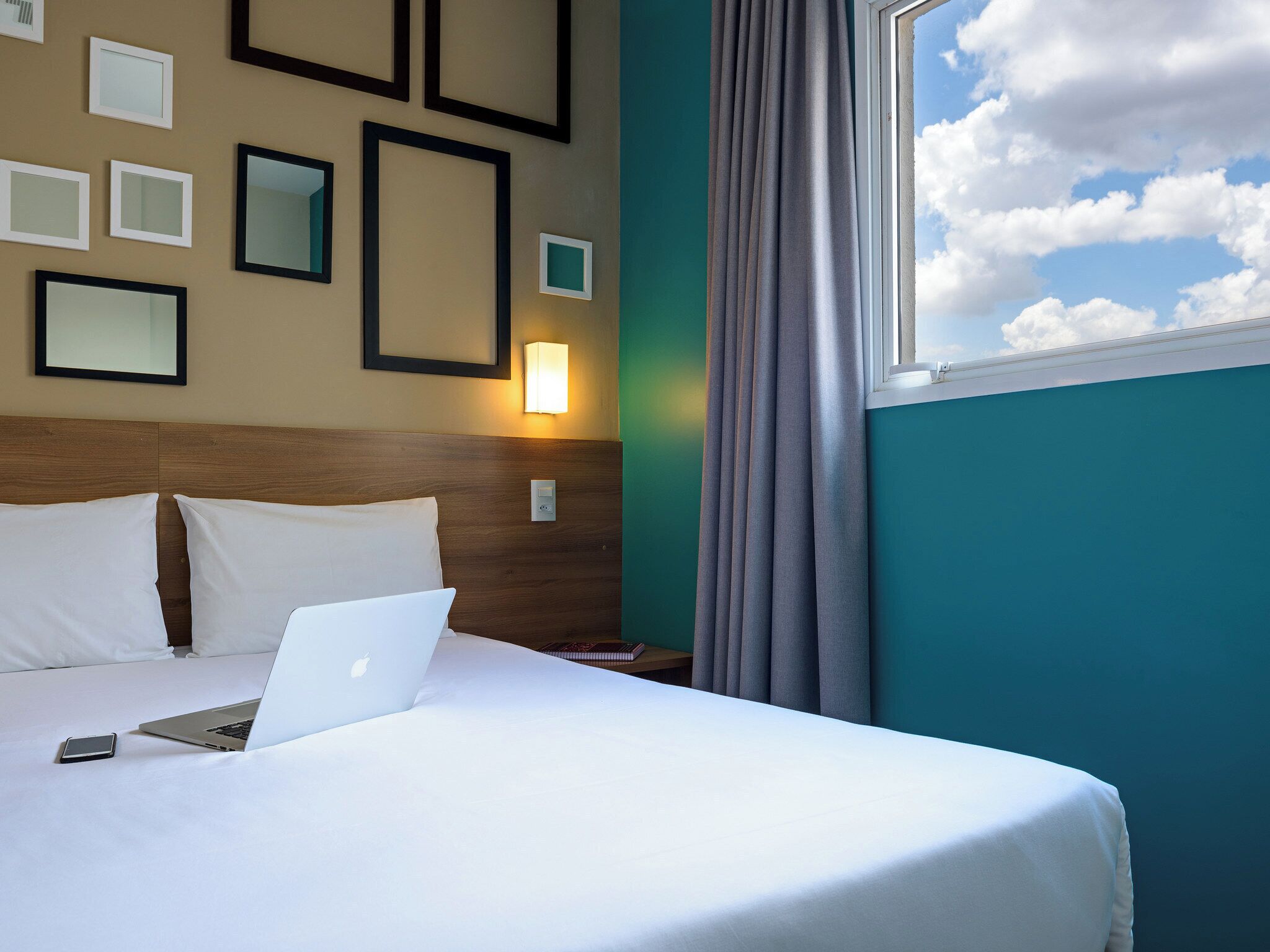 A neatly made bed with white linens, a laptop, and a smartphone on the bed. There are framed pictures on the wall and a wall-mounted light fixture.