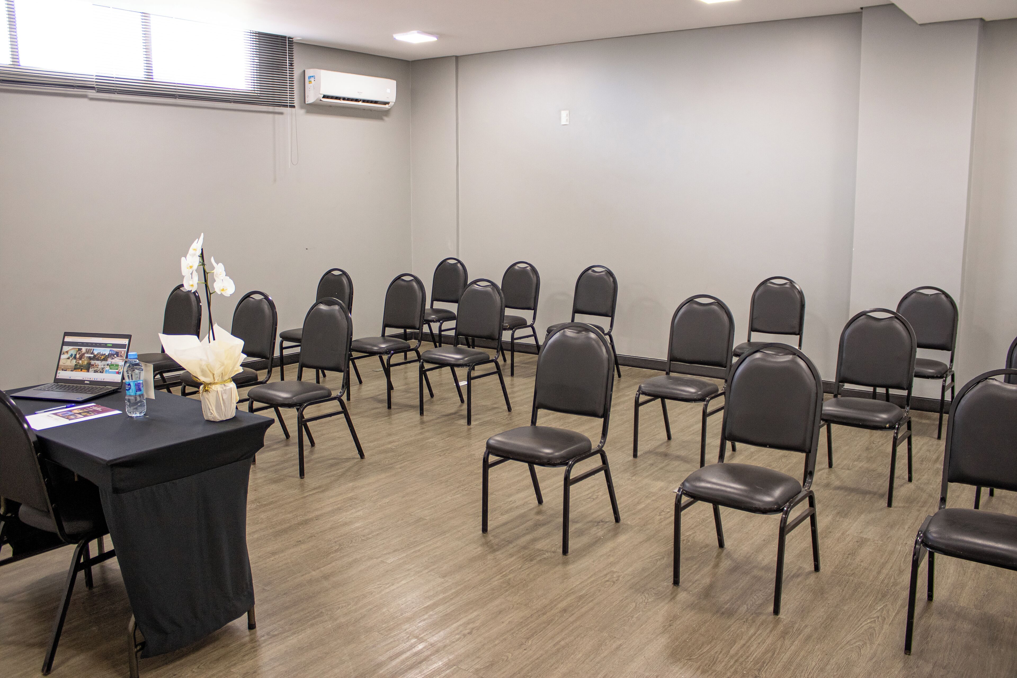Uma sala de conferências com fileiras de cadeiras, uma mesa com um laptop e uma garrafa de água, e um aparelho de ar condicionado na parede.