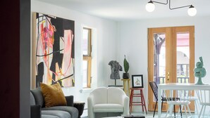 A modern living room with a sofa, armchair, coffee table, and ottoman. There is a large abstract painting on the wall, a dining table with chairs, and a staircase leading to an upper level. - The Trail in Adams - Berkshires’ Art Hotel (Adams)