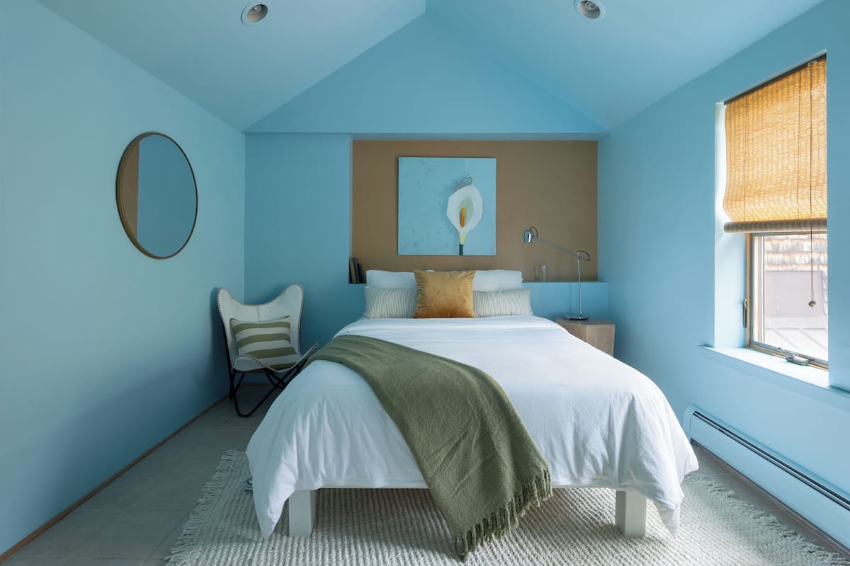 A bedroom with a bed, a chair, a bedside table, a lamp, a window with blinds, and a painting on the wall.