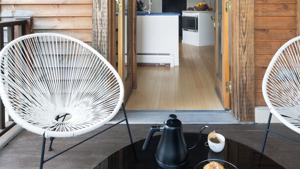 A wooden cabin interior with a dining area featuring two white chairs, a table with a coffee pot and a cup, and a view into the kitchen through an open doorway. - The Trail in Adams - Berkshires’ Art Hotel (Adams)