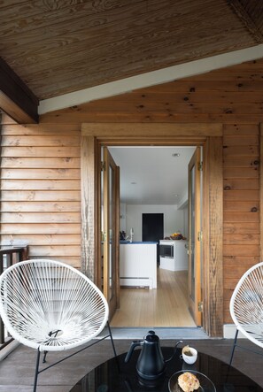 A wooden cabin interior with a dining area featuring two white chairs, a table with a coffee pot and a cup, and a view into the kitchen through an open doorway.