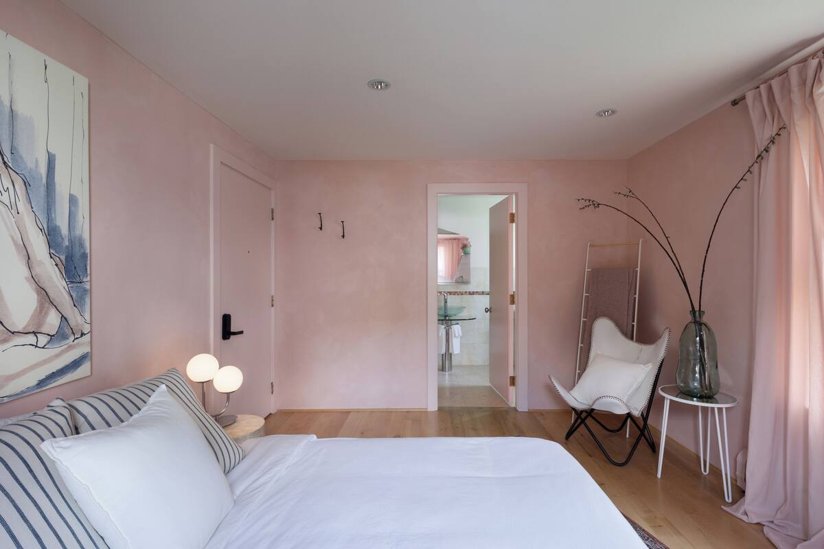 A bedroom with a bed, bedside table, and a chair. There is a door leading to a bathroom.