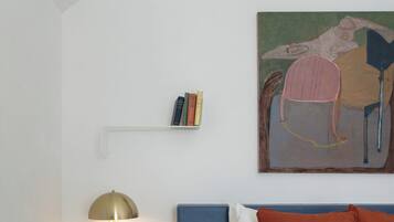 A bedroom with a bed, a bedside table with a lamp, a small shelf with books, and a painting on the wall.