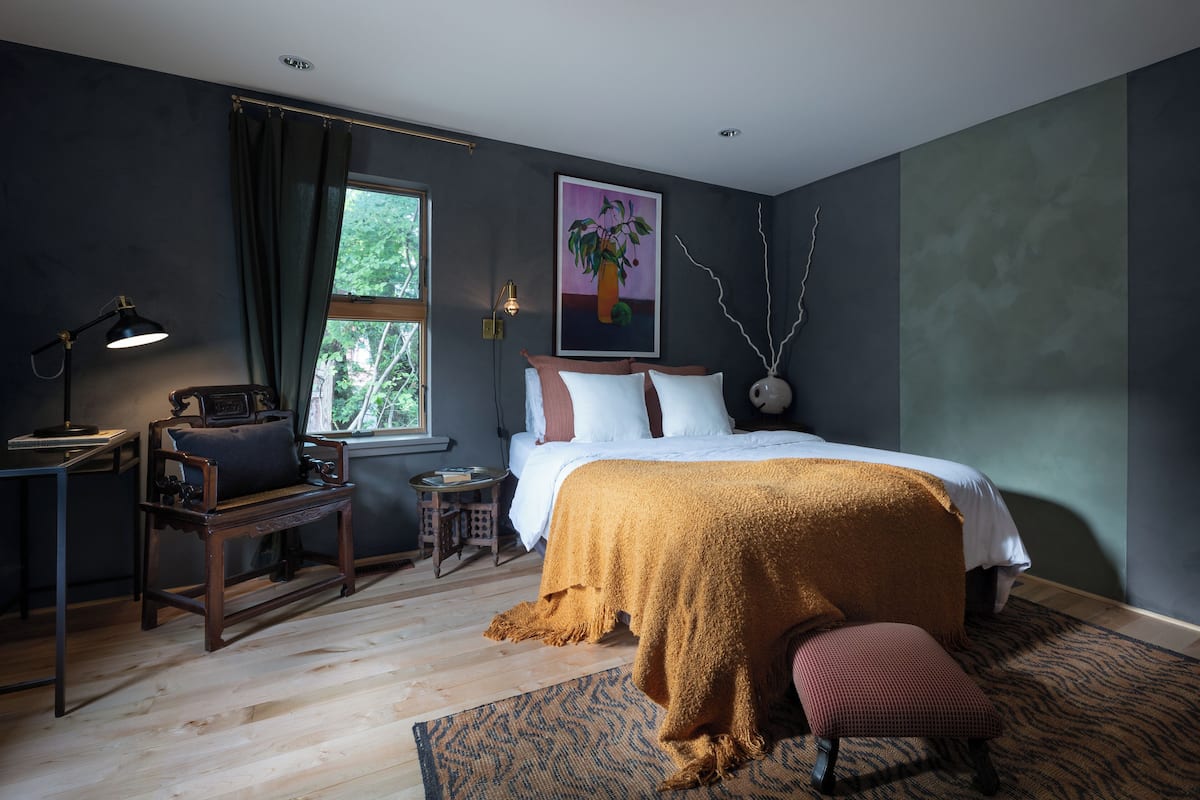 A bedroom with a bed, a desk, a chair, a window, and a painting on the wall.