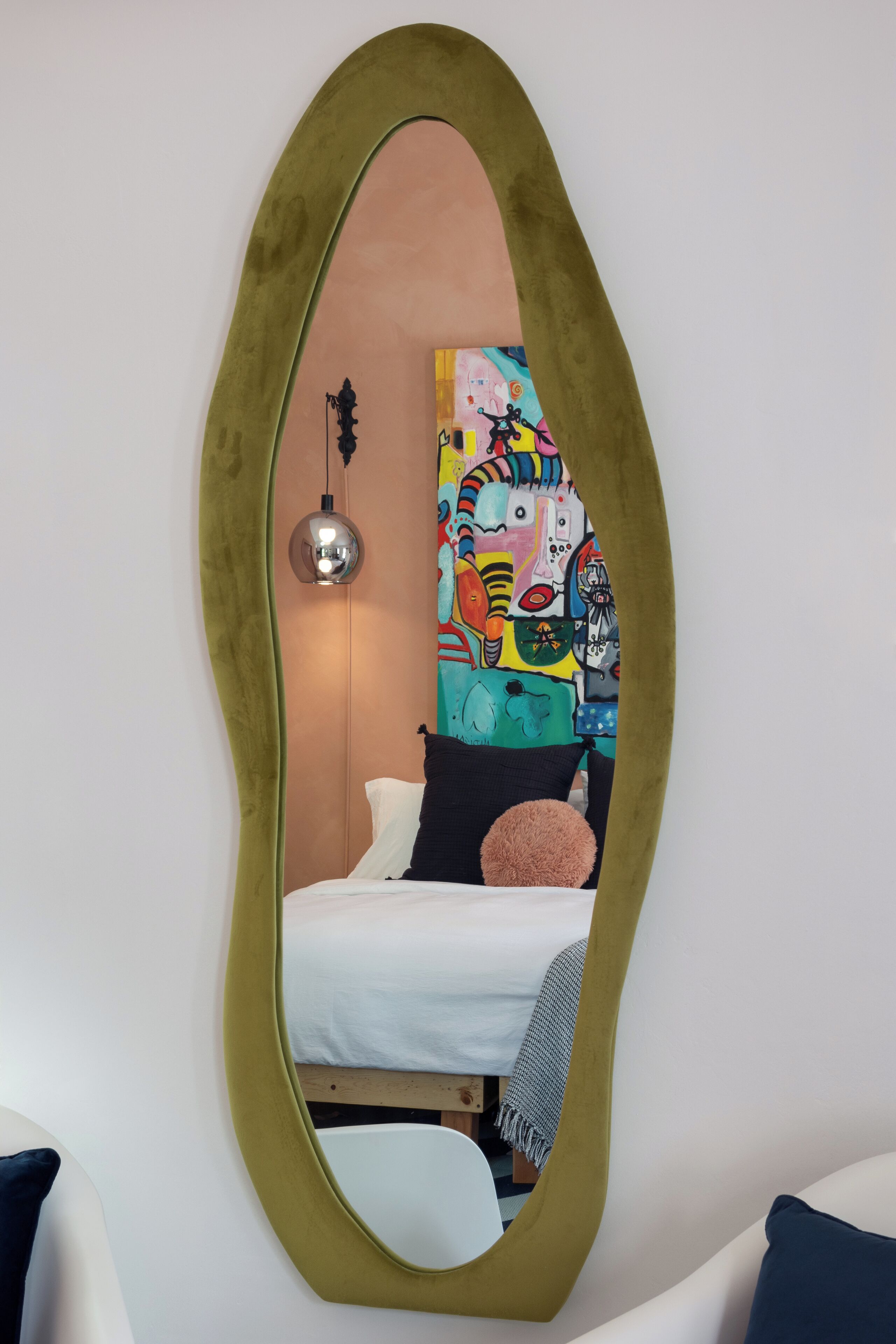 A room with a large oval mirror, a bed with white bedding, a colorful abstract painting on the wall, and a hanging lamp.