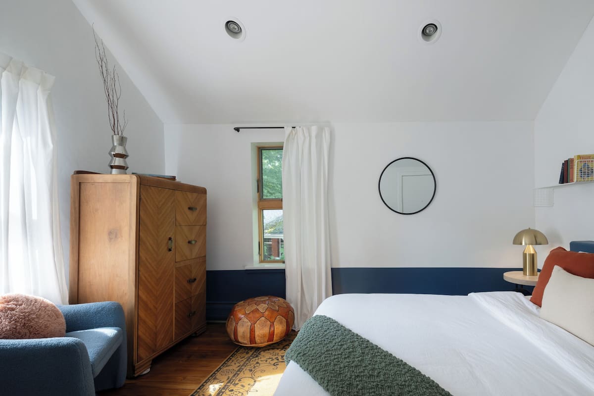 A bedroom with a bed, a wooden wardrobe, a blue armchair, a round ottoman, and a painting on the wall.
