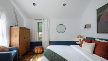 A bedroom with a bed, a wooden wardrobe, a blue armchair, a round ottoman, and a painting on the wall.