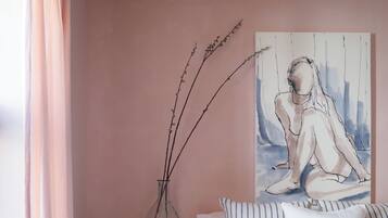 A bedroom with a bed, a vase with branches, a painting, and a round stool.