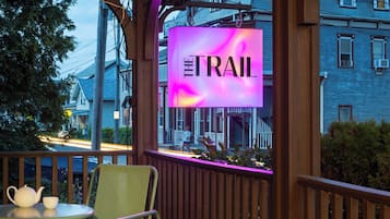 Une terrasse en bois avec une table et des chaises, un panneau lumineux au néon indiquant « THE TRAIL », et une vue sur des maisons en arrière-plan.