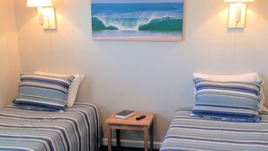 A hotel room with two beds, each with striped bedding, a small wooden nightstand with a book and remote control, and a painting of a wave on the wall. - Village Marina (Hatteras)
