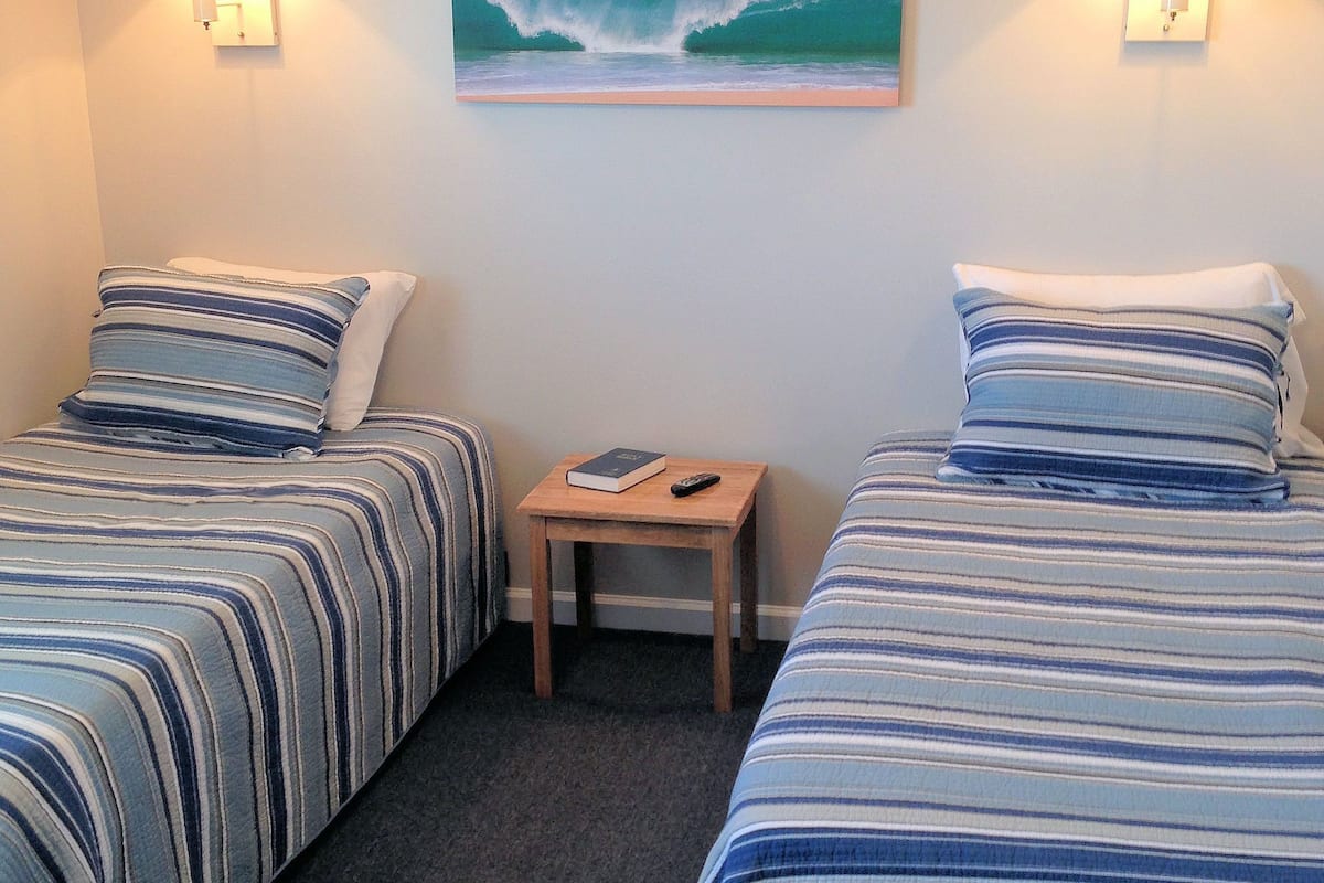 A hotel room with two beds, each with striped bedding, a small wooden nightstand with a book and remote control, and a painting of a wave on the wall.