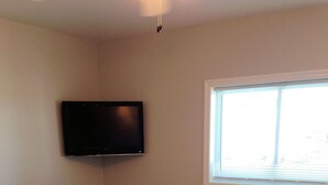 A dining area with a round table and four chairs, a flat-screen TV mounted on the wall, a window with blinds, and a wall-mounted air conditioning unit. - Village Marina (Hatteras)