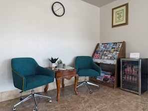 A room with two teal chairs, a wooden table with a plant, a clock, a framed picture, a magazine rack, and a refrigerator with soda cans. - Budget Inn By Hotel O Ada OK (Ada)
