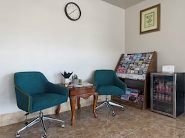 Una habitación con dos sillas color turquesa, una mesa de madera con una planta, un reloj, un cuadro enmarcado, un mueble para revistas y un refrigerador con latas de refresco.