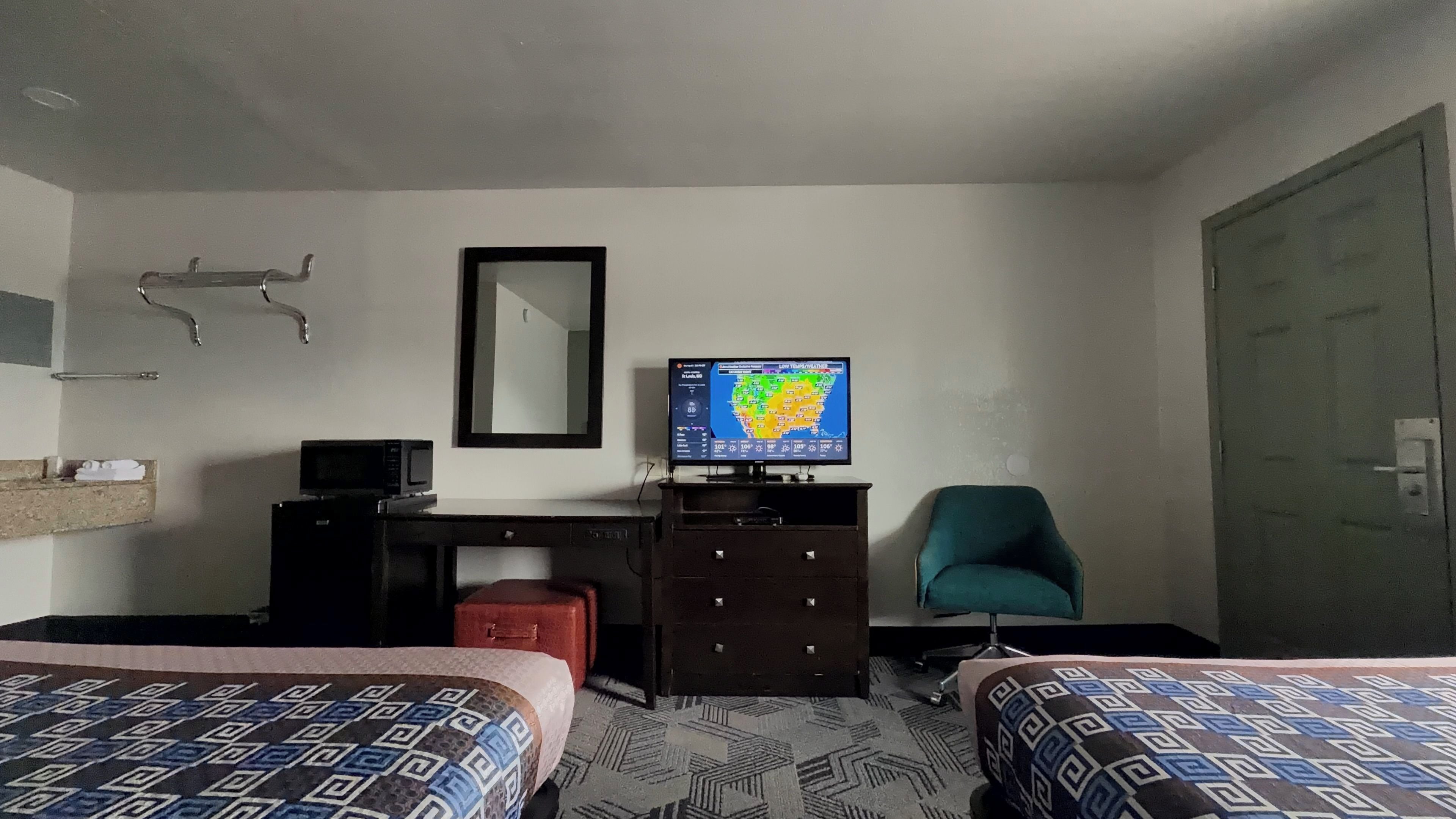 A hotel room with two beds, a desk with a computer, a chair, a TV displaying a weather map, and a bathroom with a towel rack.