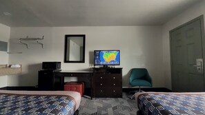 Habitación de hotel con dos camas, un escritorio con computadora, una silla, un televisor mostrando un mapa del tiempo y un baño con perchero.