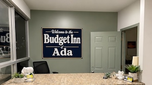 A reception desk with a sign reading 'Welcome to the Budget Inn of Ada,' a potted plant, a small statue, and a door in the background. - Budget Inn (Ada)