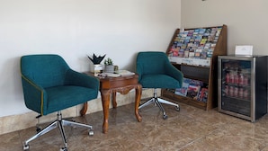Two teal chairs, a wooden table, a clock, a framed picture, a magazine rack, and a refrigerator with soda cans. - Budget Inn (Ada)