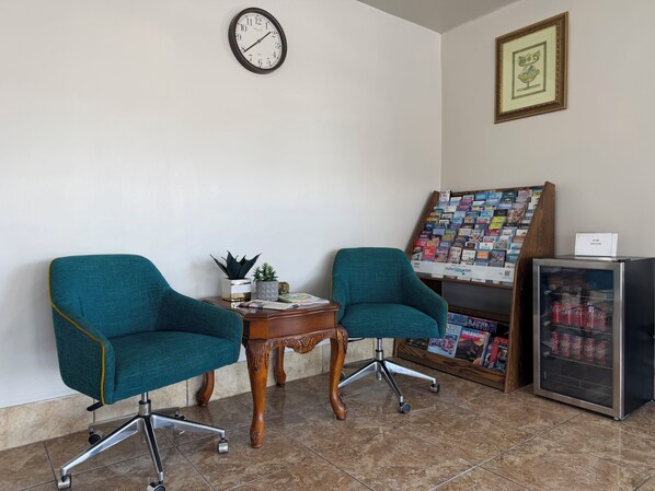 Two teal chairs, a wooden table, a clock, a framed picture, a magazine rack, and a refrigerator with soda cans. - Budget Inn (Ada)