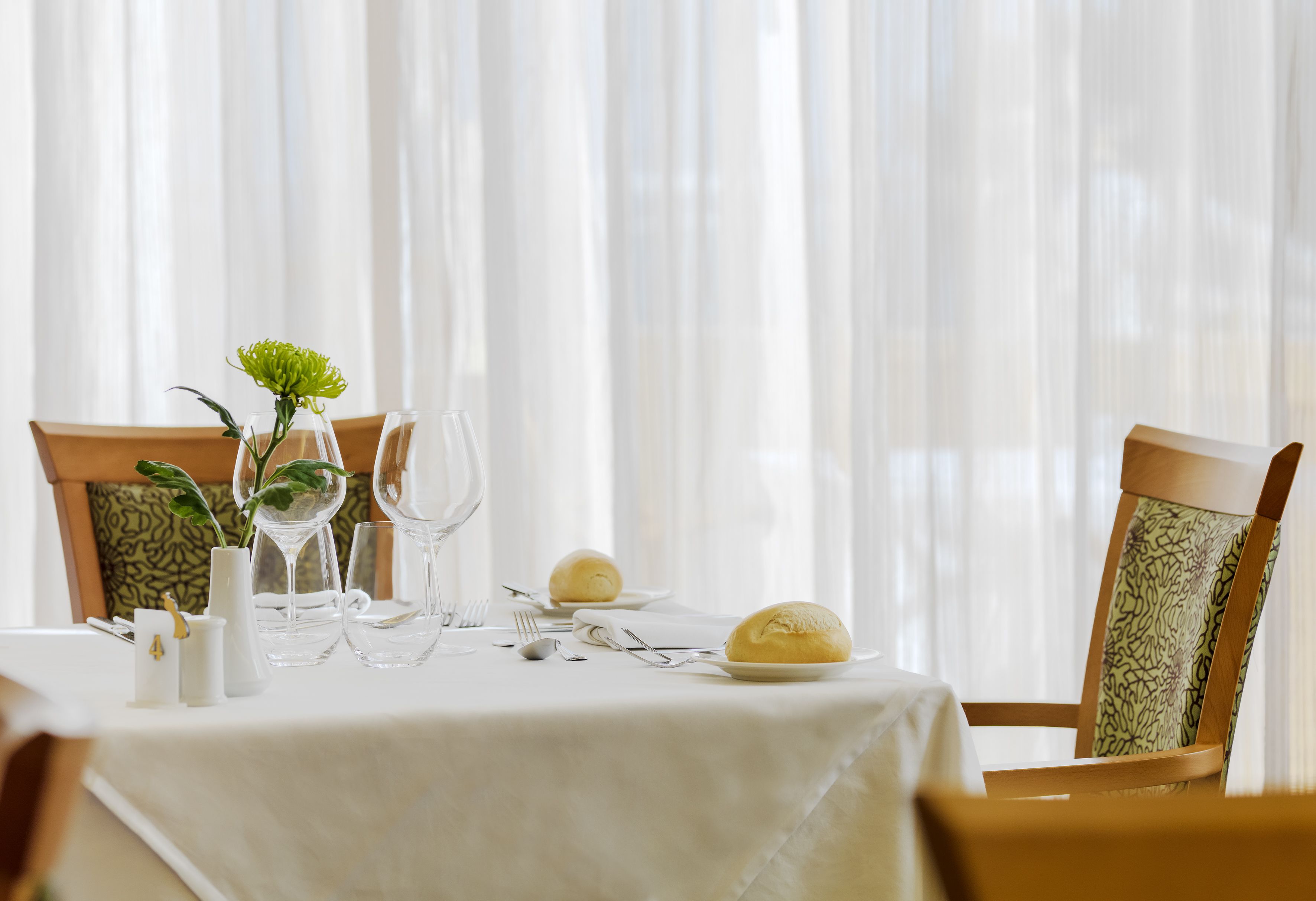 Une table à manger dressée avec une nappe blanche, de la verrerie, des assiettes et une seule fleur dans un vase.