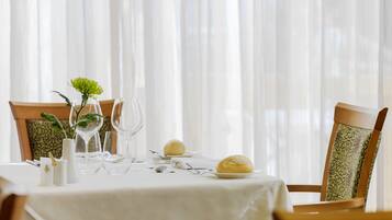 Une table à manger dressée avec une nappe blanche, de la verrerie, des assiettes et une seule fleur dans un vase.