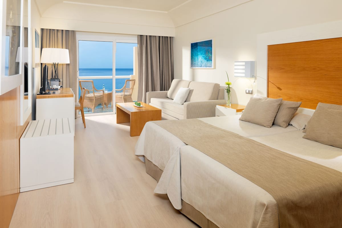 A hotel room with a bed, a desk, a chair, a sofa, a coffee table, and a view of the sea.