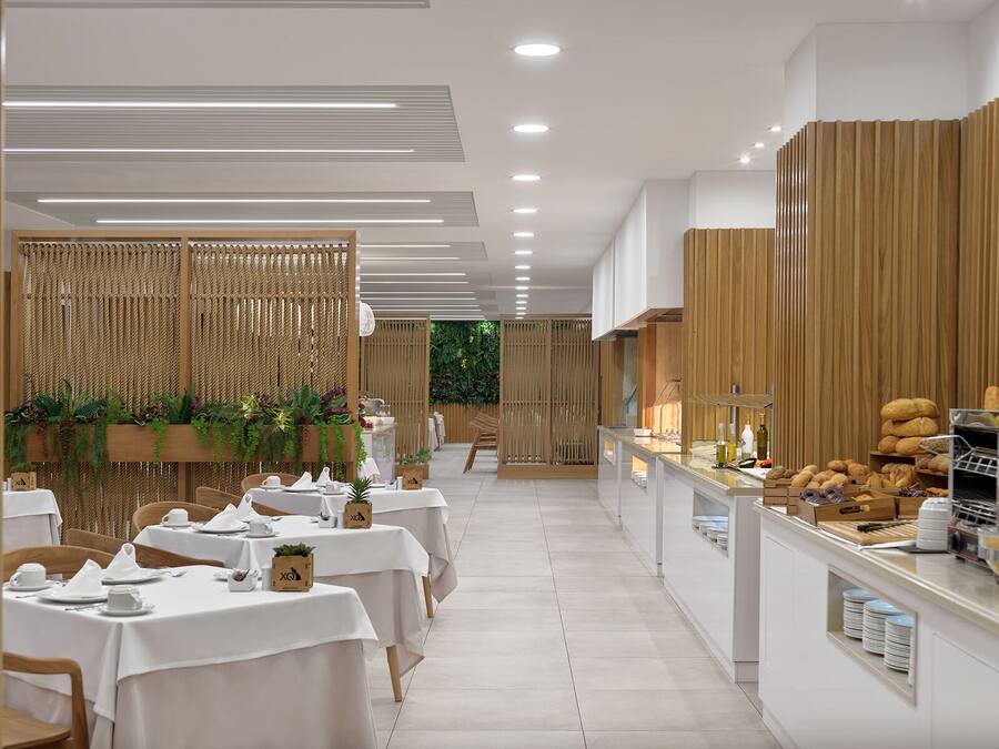 A modern restaurant with white tables and chairs, a buffet counter with bread and pastries, and vertical wooden panels.