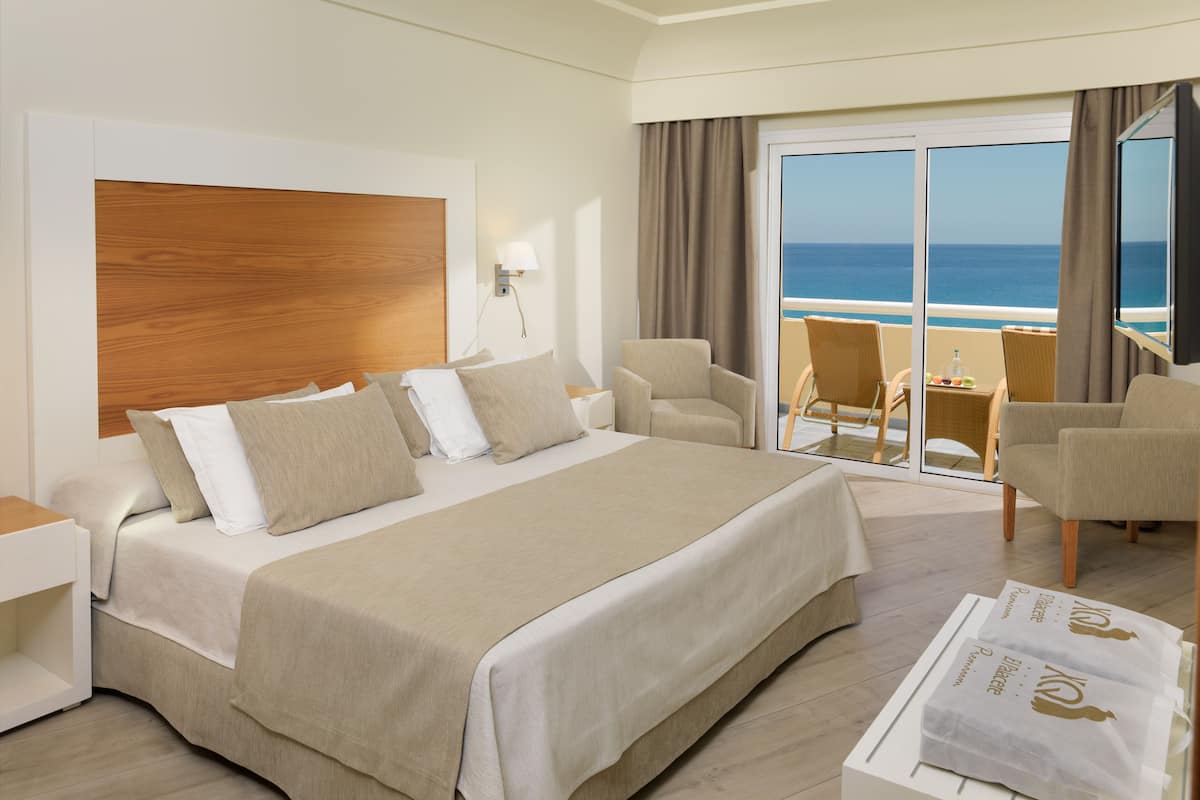 A hotel room with a large bed, two armchairs, a small table, and a view of the ocean through a sliding glass door.