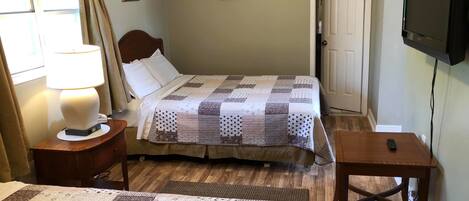 Quarto com duas camas, uma mesinha de cabeceira com abajur, televisão na parede e uma porta.