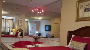 A hotel room with a large bed, a bedside table, a mirror, a television, and a bathroom visible in the reflection. - Niagara Inn & Suites (Niagara Falls)