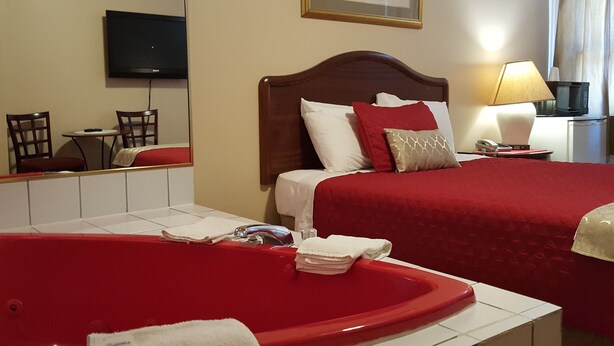 A hotel room with a large red bathtub, a wooden bed with red bedding, a television on the wall, and a small desk with a lamp. - Niagara Inn & Suites (Niagara Falls)
