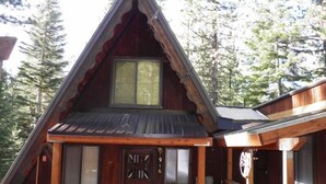 A wooden cabin with a steeply pitched roof, a front door with a window, and a porch area. - 916 Kiowa (South Lake Tahoe)