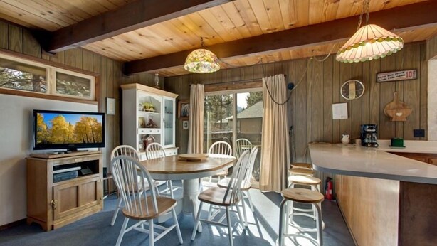 A cozy cabin interior with a wooden dining table, white chairs, a bar area, and a television on a wooden cabinet. - 890 Candlewood Drive (South Lake Tahoe)